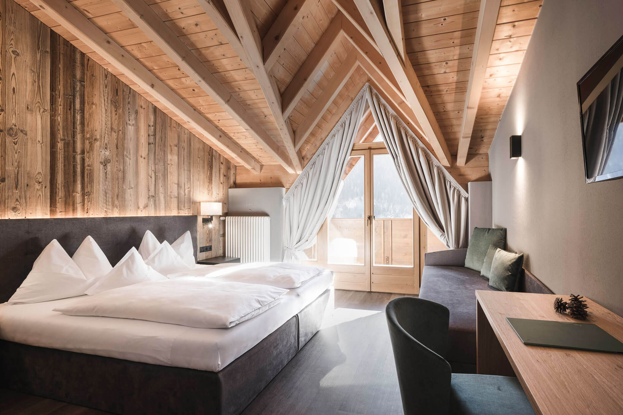 Großes Hotelzimmer mit Doppelbett, grauer Couch, Schreibtisch und Blick auf den Balkon - Hotel Alpenhof