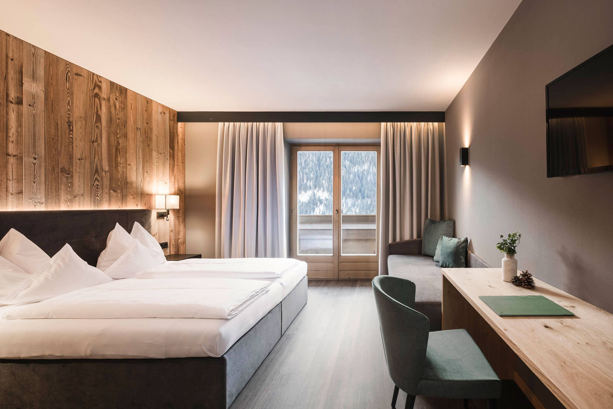 Doppelbett im Zimmer Seegruben mit grauer Couch, Schreibtisch und Blick auf den Balkon - HOtel Alpenhof