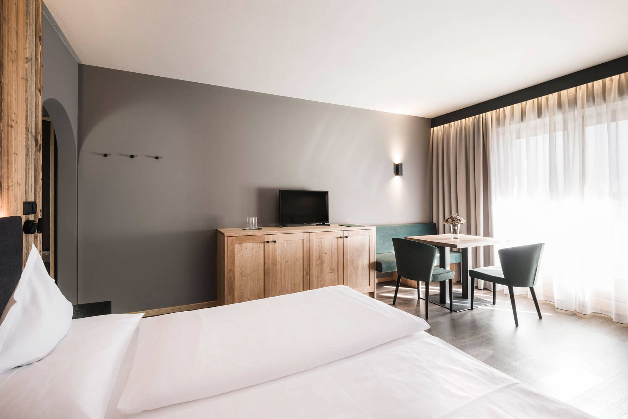 Das Spitzen Zimmer mit Doppelbett, Schrank, darüber ein Fernseher und daneben eine kleine Sitzecke - Hotel Alpenhof